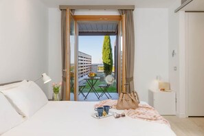 Een hotelkamer met een bed, nachtkastje, lamp, uitzicht op een balkon met tafel en stoelen, en een hoge boom.