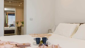 A hotel room with a bed, a bedside table with two mugs, a tray with a handbag, and a view of the outside.