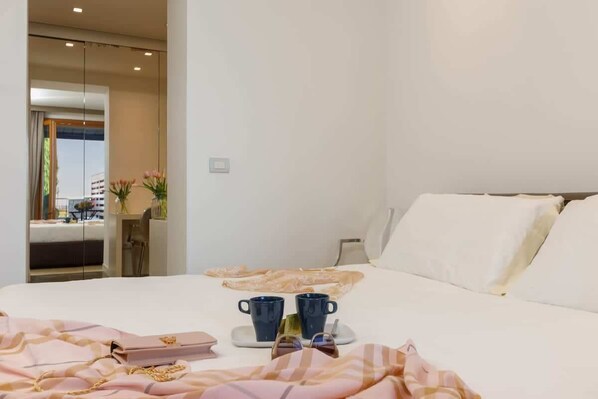 A hotel room with a bed, a bedside table with two mugs, a tray with a handbag, and a view of the outside.