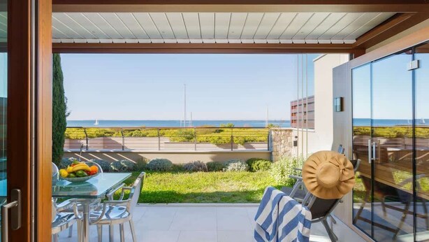 A balcony with a glass door, a table with chairs, a hat, and a view of the sea.