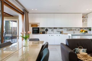 Een moderne keuken met witte kastjes, een eethoek met een glazen tafel, en een zwarte bank.
