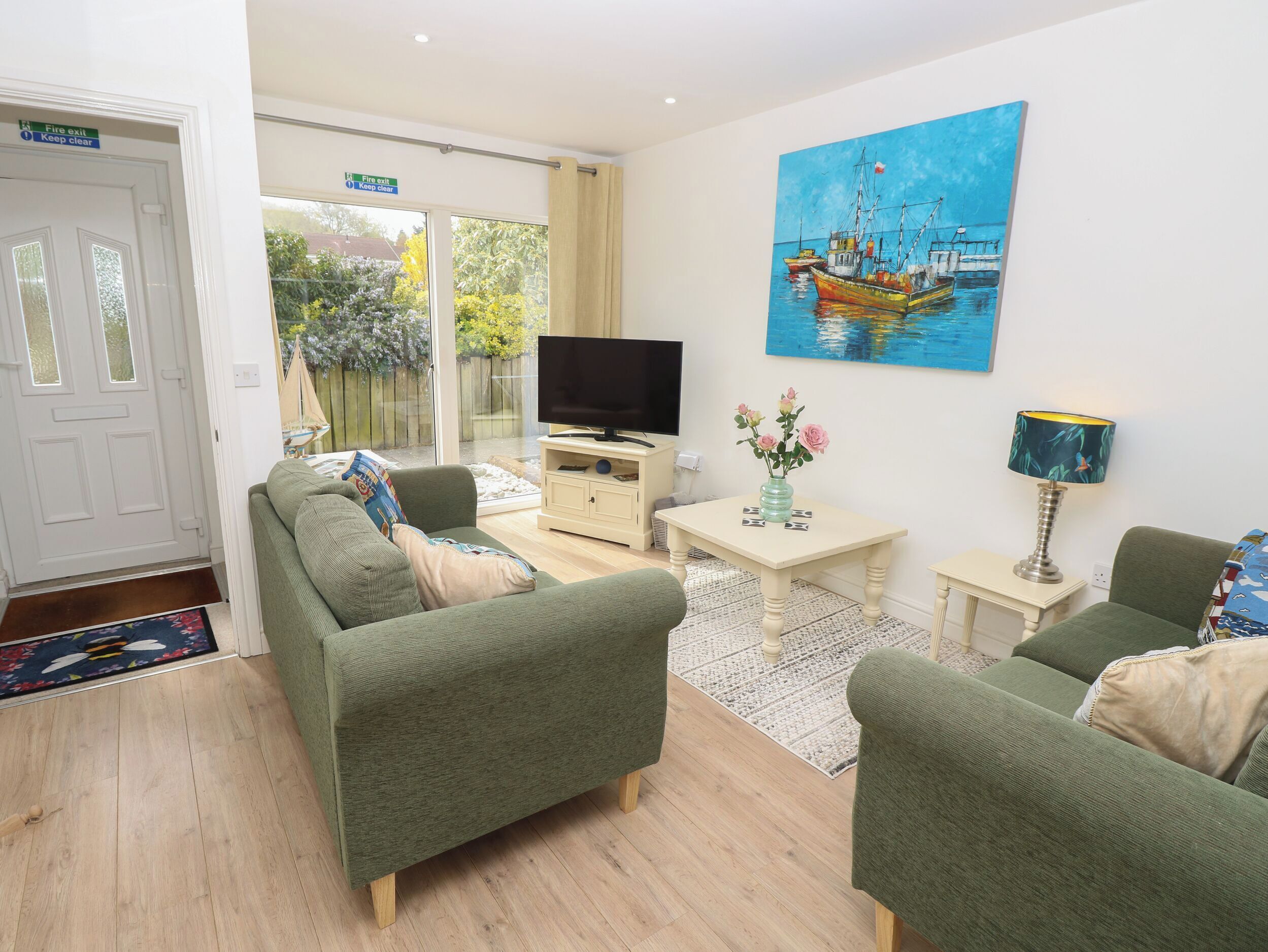 A living room with a green sofa, a TV, a painting of boats, a vase with flowers, and a lamp.