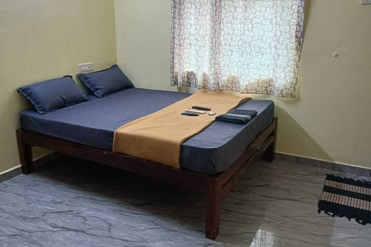 Un lit simple avec un cadre en bois, une literie bleue et une couverture marron. On y trouve deux oreillers, une télécommande et un smartphone. Un rideau à motifs est partiellement tiré, et un tapis rayé est visible sur le sol.