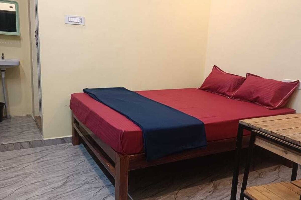 Une petite chambre avec un lit simple, un cadre de lit en bois, un matelas rouge et une couverture bleue. Une petite table en bois se trouve à côté du lit. Un lavabo et un miroir sont visibles dans le coin de la pièce.