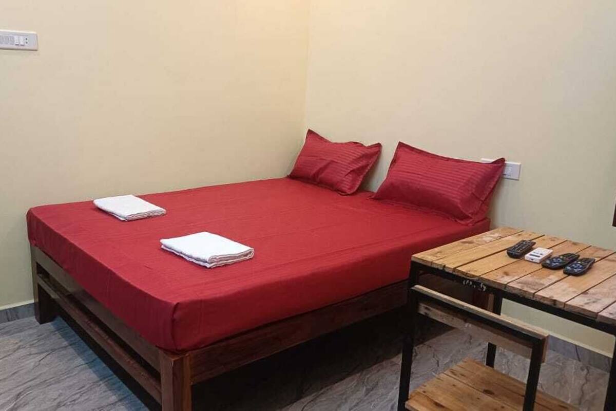 Un lit simple avec une literie rouge, une table de chevet en bois avec une lampe et une petite table avec des télécommandes.