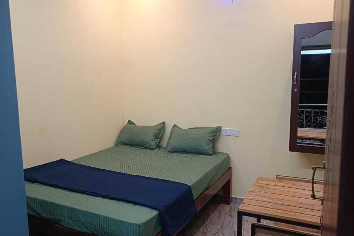 Une petite chambre avec un lit simple, un cadre de lit en bois, un matelas vert et des oreillers assortis. Il y a une petite table en bois à côté du lit et une fenêtre avec une grille en métal.