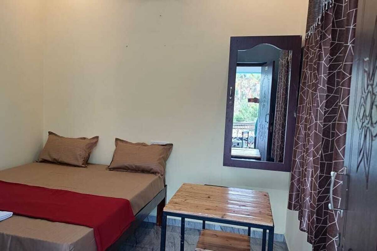 Un lit simple avec une couverture rouge, deux oreillers et une petite table en bois placée à côté.