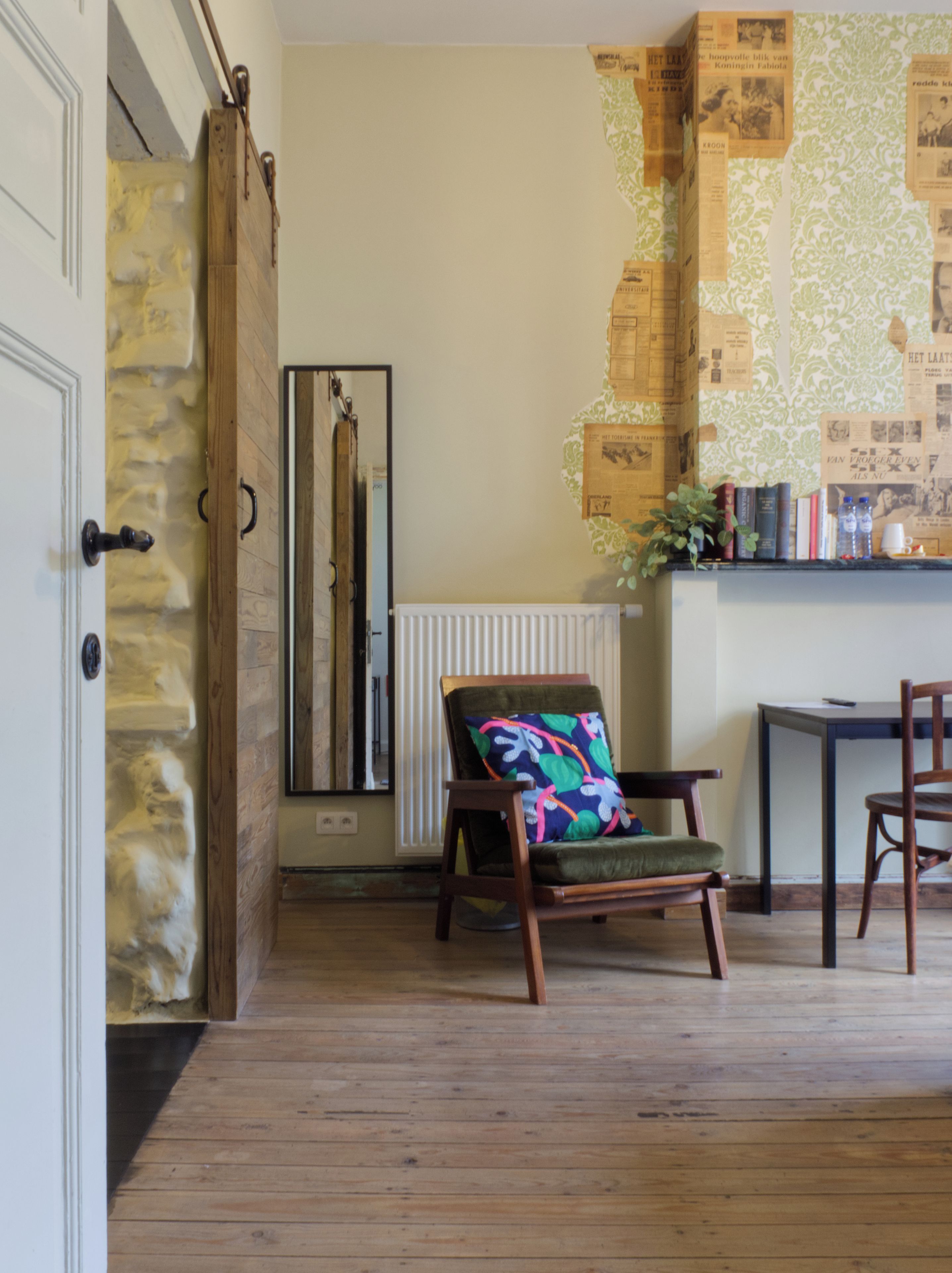 Une pièce avec un plancher en bois, une chaise dotée d’un coussin coloré, un radiateur et un mur recouvert de coupures de journaux.