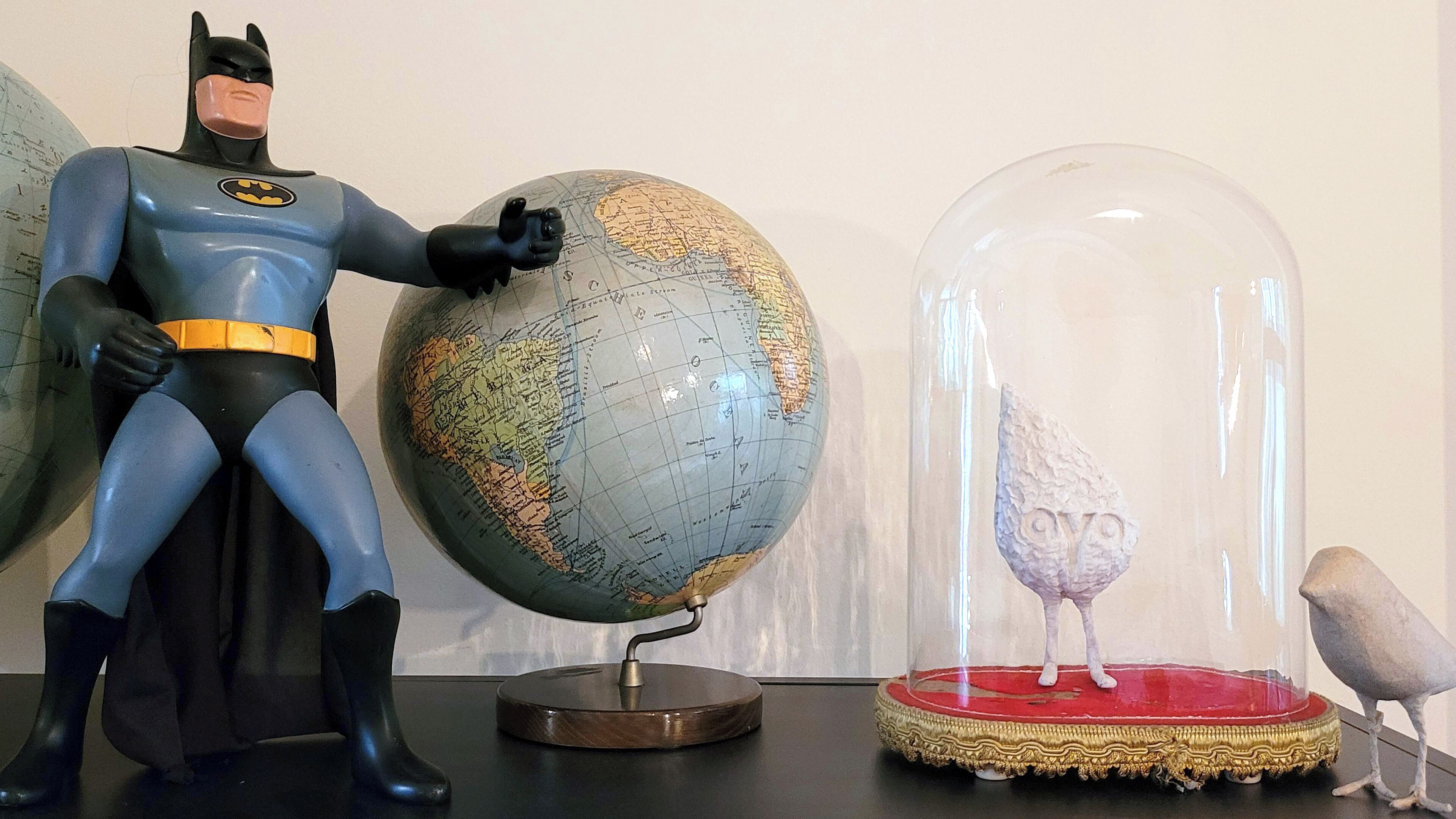 Una figura de acción de Batman, un globo terráqueo y una figurita de pájaro dentro de una cúpula de vidrio.