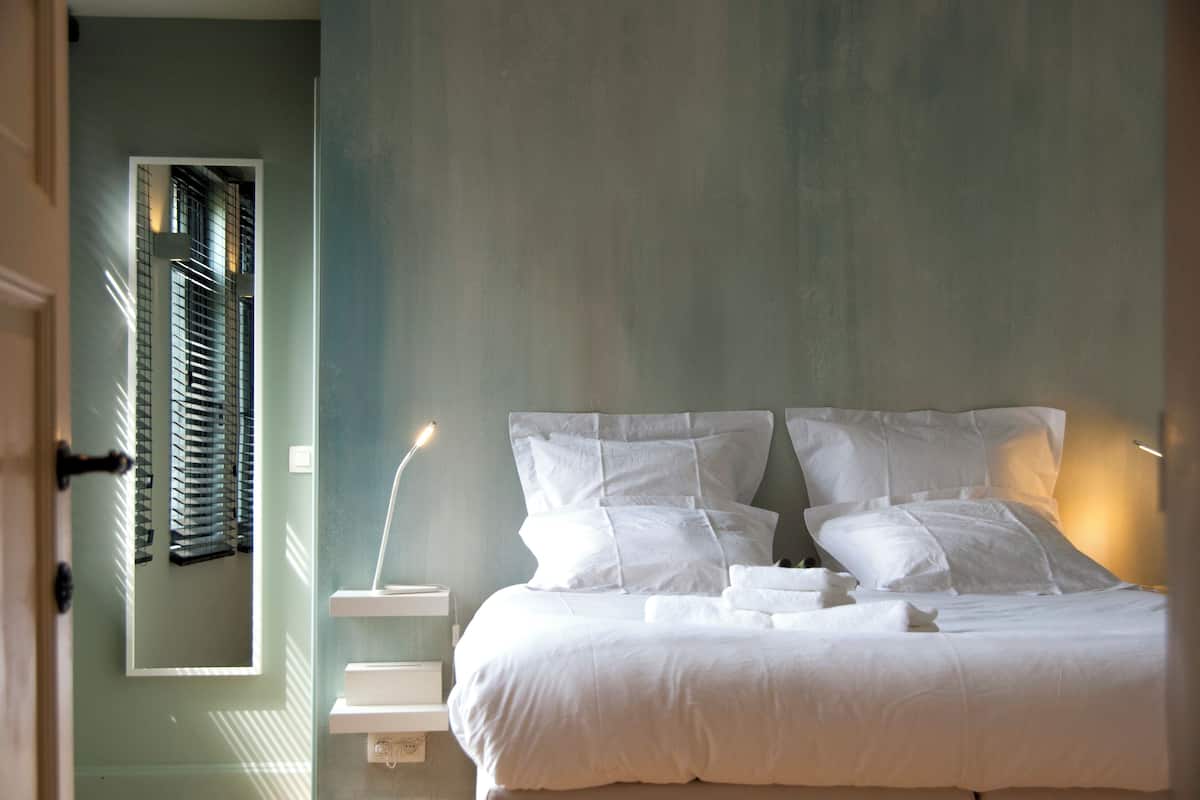 En pænt redt seng med hvide sengetøj og puder, et natbord med en lampe og et vindue med persienner.