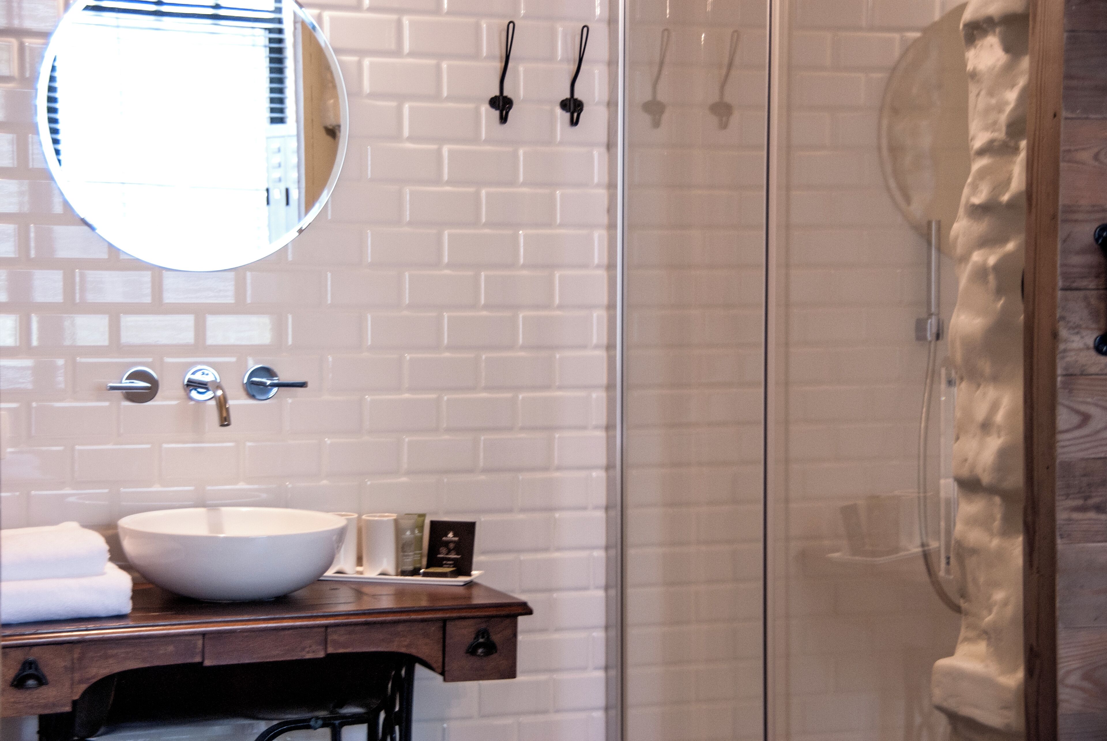 A bathroom with a white tiled wall, a round mirror, a wooden vanity with a white sink, and a glass shower enclosure.