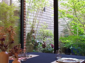 A dining table set with a vase of dried flowers, a glass, and a plate, with a view of a garden through a window. - OYO  (Kortrijk)