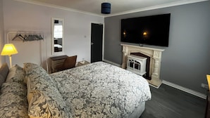 Une chambre dotée d’un grand lit, d’un téléviseur à écran plat fixé au-dessus d’une cheminée, d’un bureau avec une chaise et d’un luminaire suspendu.