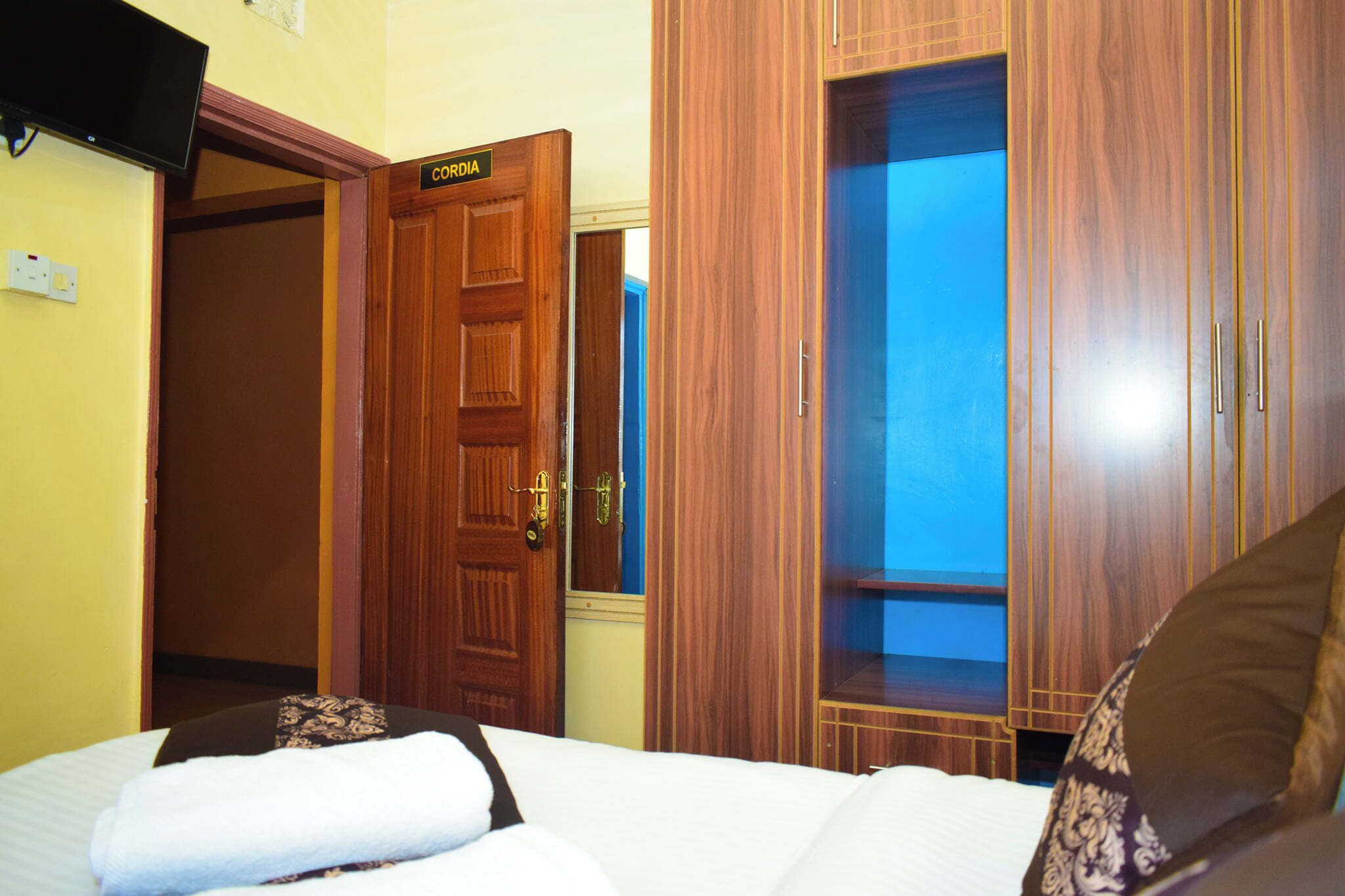 A hotel room with a bed, a wooden wardrobe, a closed door labeled "CORDIA," and a television mounted on the wall.