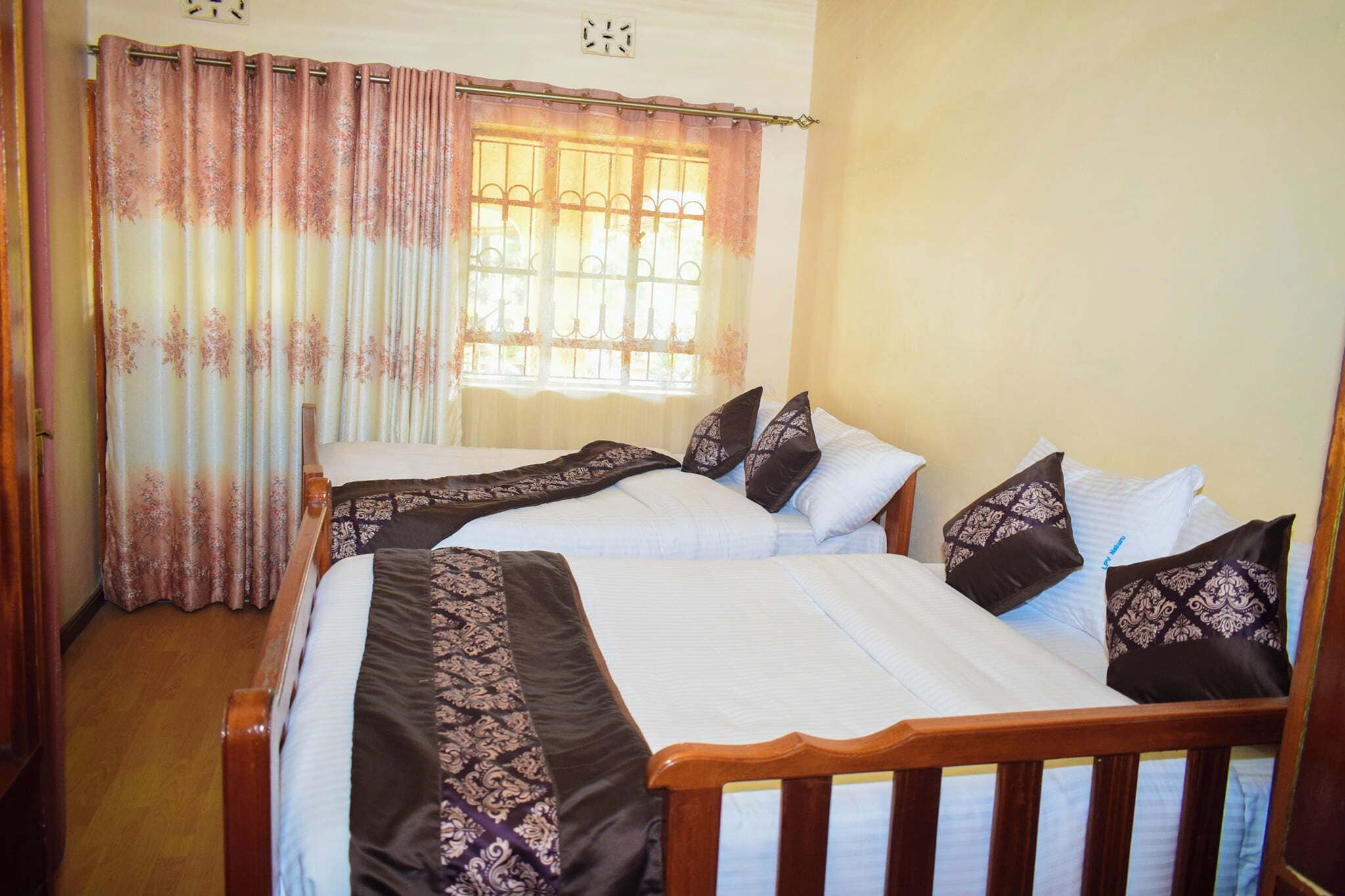 A room with two beds, each with a wooden frame and white bedding. There is a window with bars and curtains, and a clock on the wall.
