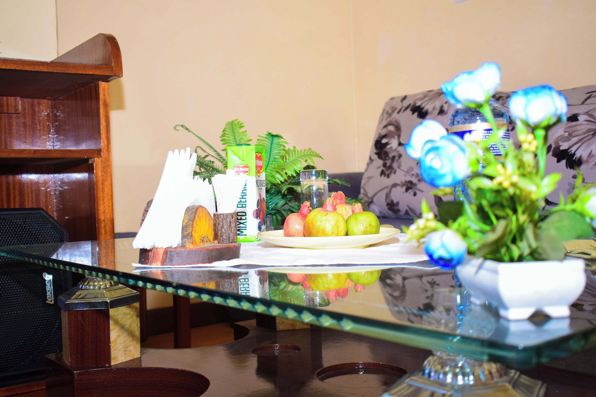 A dining table with a glass top, surrounded by a sofa, a wooden bookshelf, and a small decorative plant.