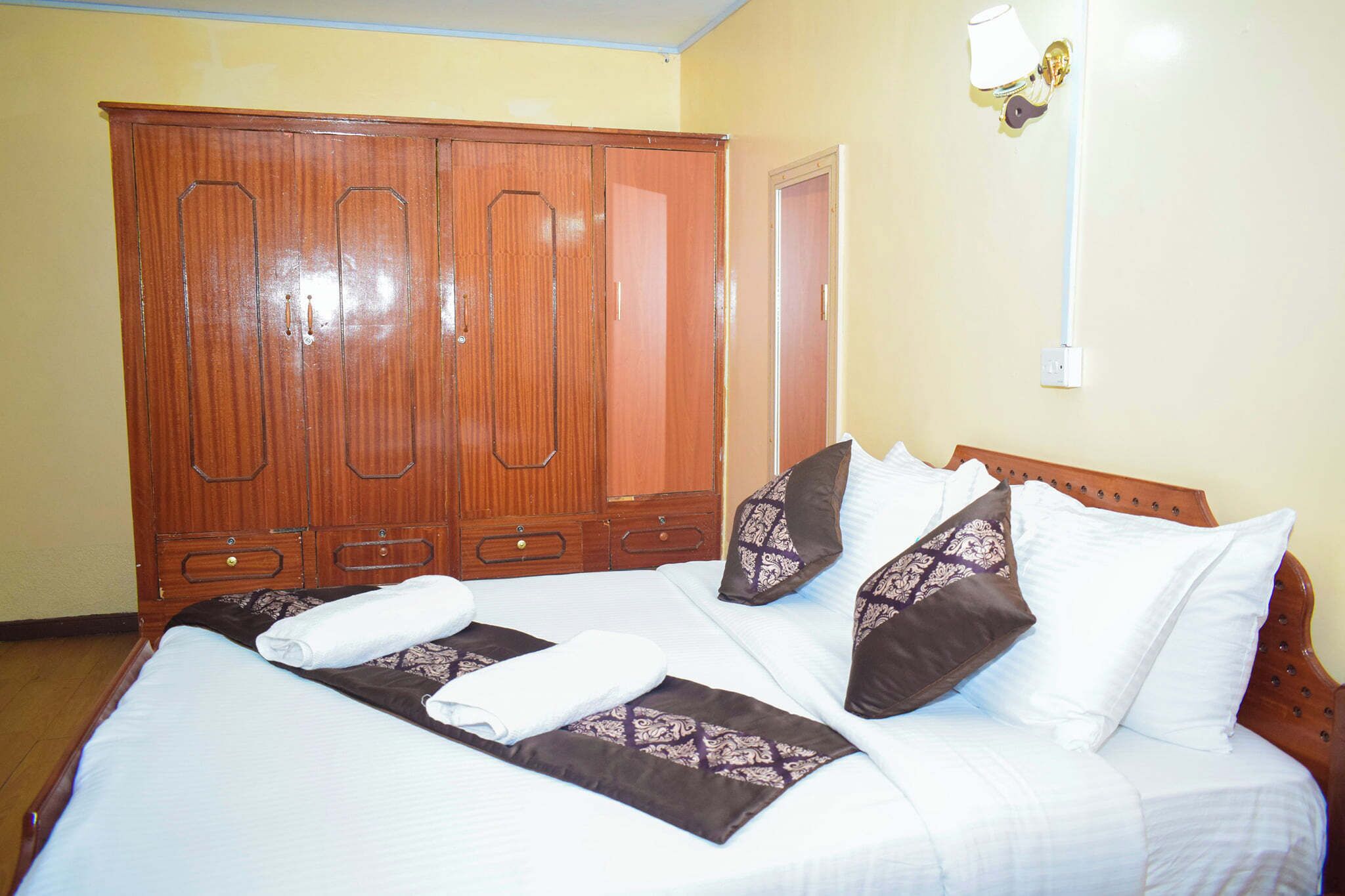 A hotel room with a large wooden wardrobe, a neatly made bed with pillows and a blanket, and a wall-mounted light fixture.
