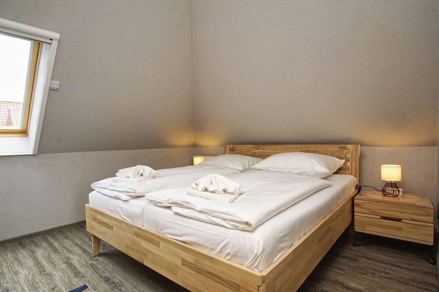 Een tweepersoonsbed met houten hoofdeinde en voeteinde, twee nachtkastjes met lampen, en een raam met zonwering.