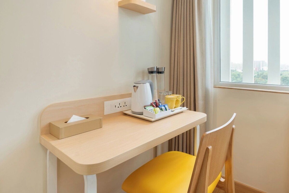 A hotel room with a desk, a chair, a coffee maker, a cup, and a window with curtains.
