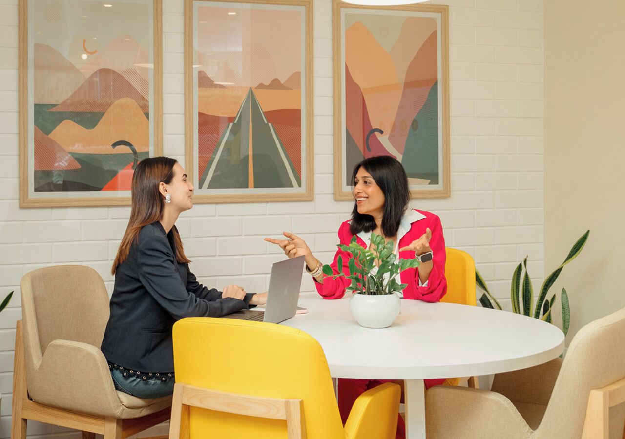 Two people sitting at a round table with a laptop, in a room with white brick walls and abstract art.