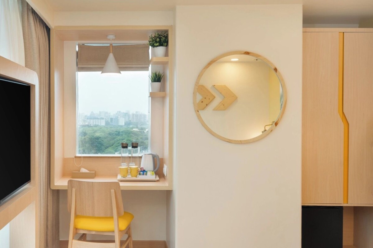 A modern room with a large flat-screen TV, a desk with a chair, a window with a city view, and a round mirror with a unique design.