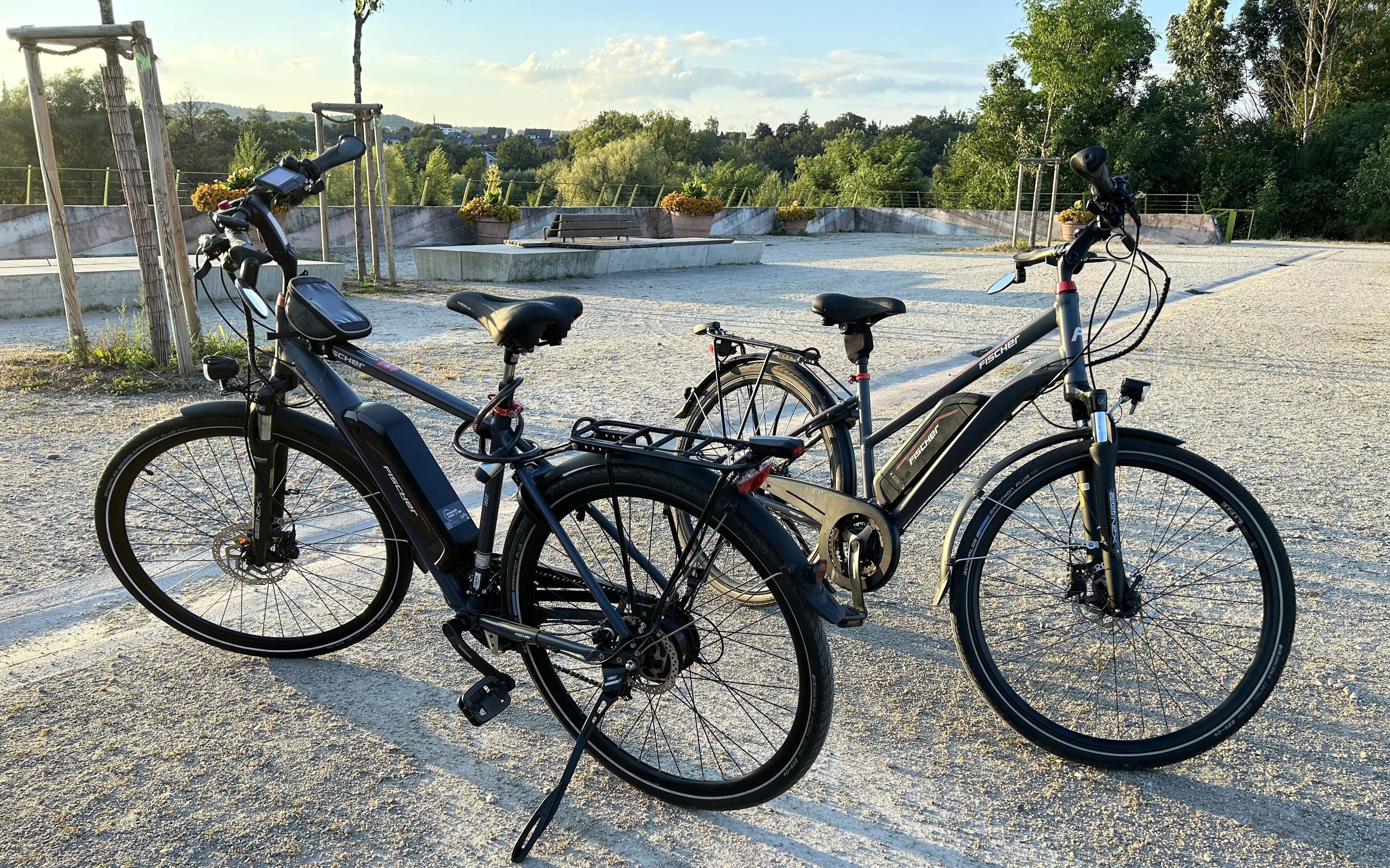 Deux vélos sont garés sur une surface de gravier, avec en arrière-plan une vue sur un parc et des bâtiments.
