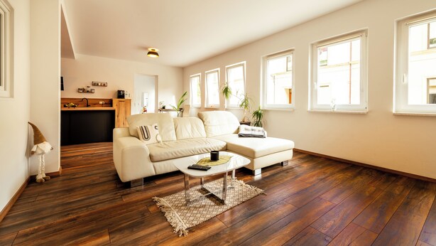Ein geräumiges Wohnzimmer mit einem weißen Ecksofa, einem Couchtisch und Holzboden. Mehrere Fenster sorgen für natürliches Licht.