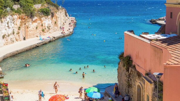 Eine Küstenlandschaft mit Strand, klarem blauen Wasser, Menschen beim Schwimmen und Sonnenbaden sowie ein Gebäude mit roter Fassade.