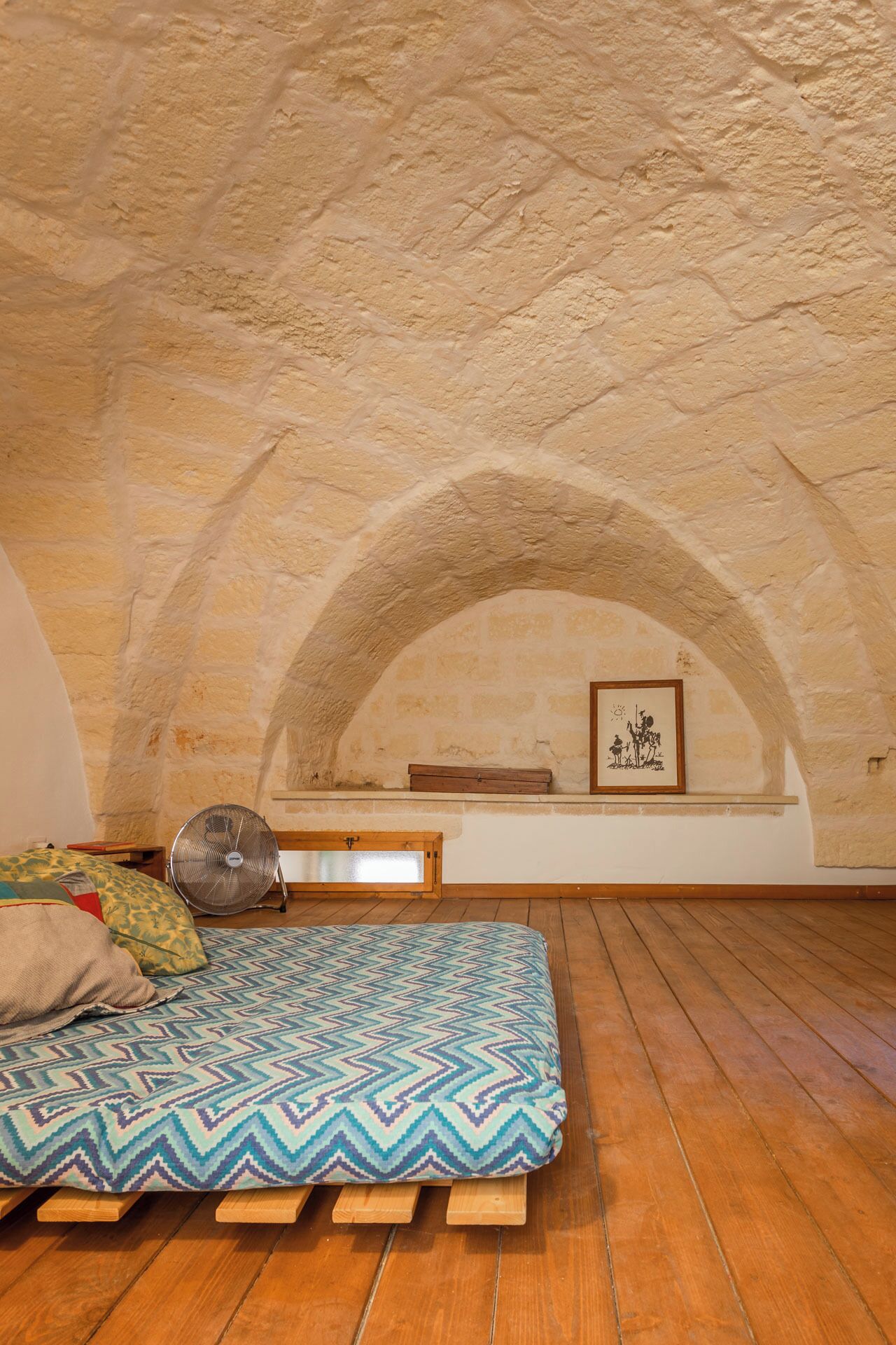 Une pièce mansardée chaleureuse avec un plancher en bois, un coussin à motifs, un ventilateur et un tableau encadré accroché au mur.