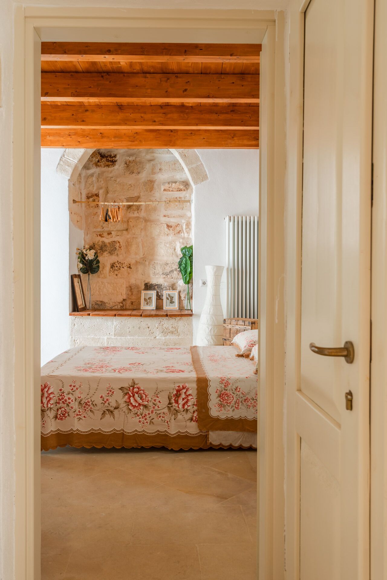An interior room with a rustic stone wall, wooden beams, a bed with floral bedding, and a white radiator.
