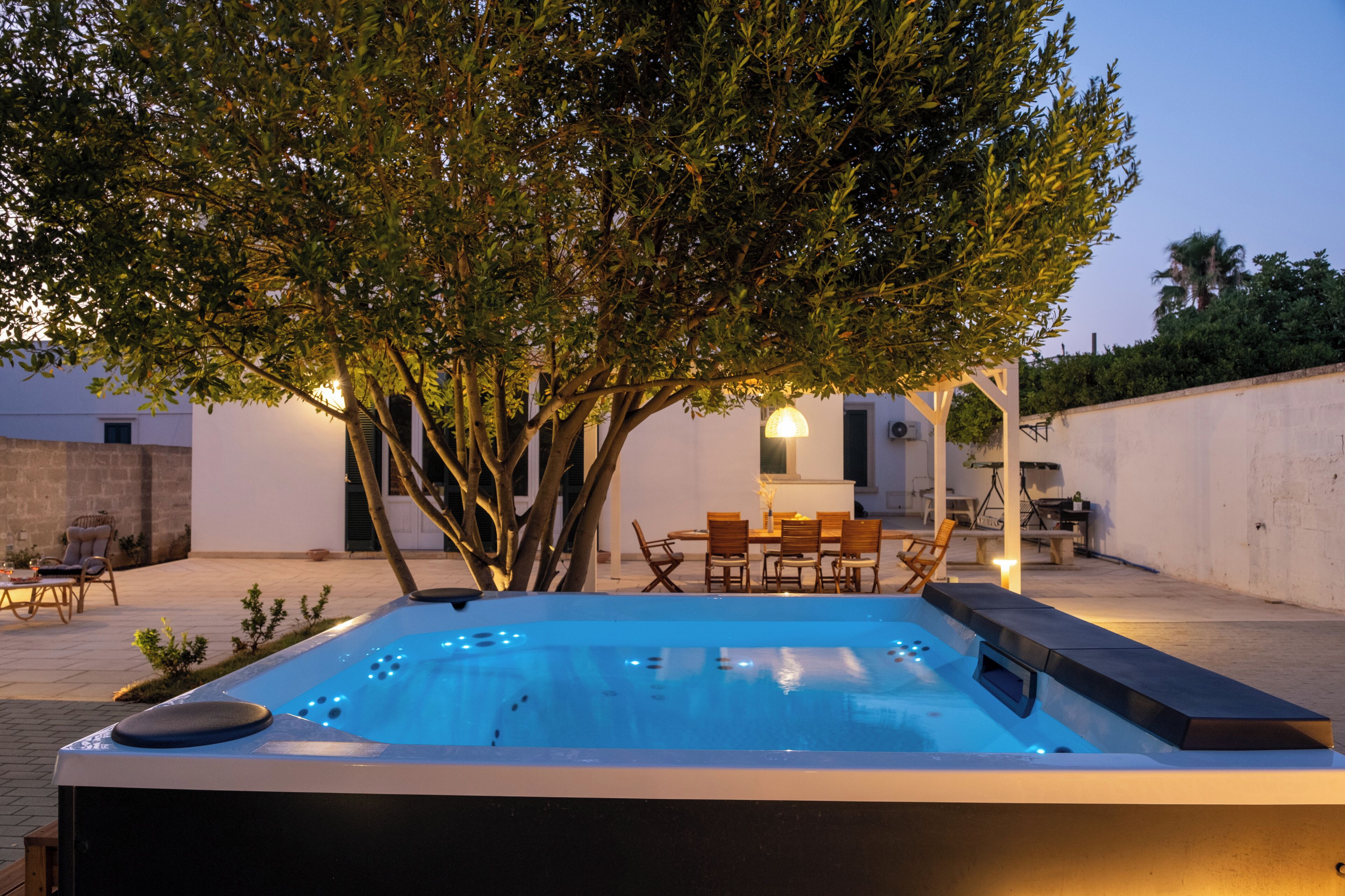 A hot tub with a tree in the background, surrounded by outdoor furniture and a white wall.