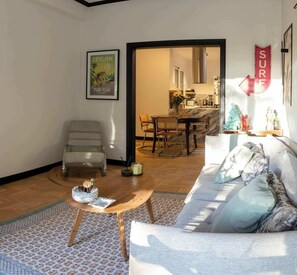 Uma sala de estar com um sofá, uma poltrona, uma mesa de centro e uma área de refeições ao fundo.