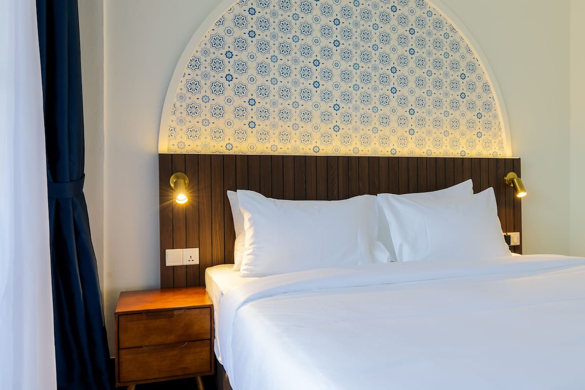 Un lit bien fait, avec du linge de lit blanc, une table de chevet en bois dotée d’un tiroir, et un mur orné d’un motif décoratif au-dessus du lit.