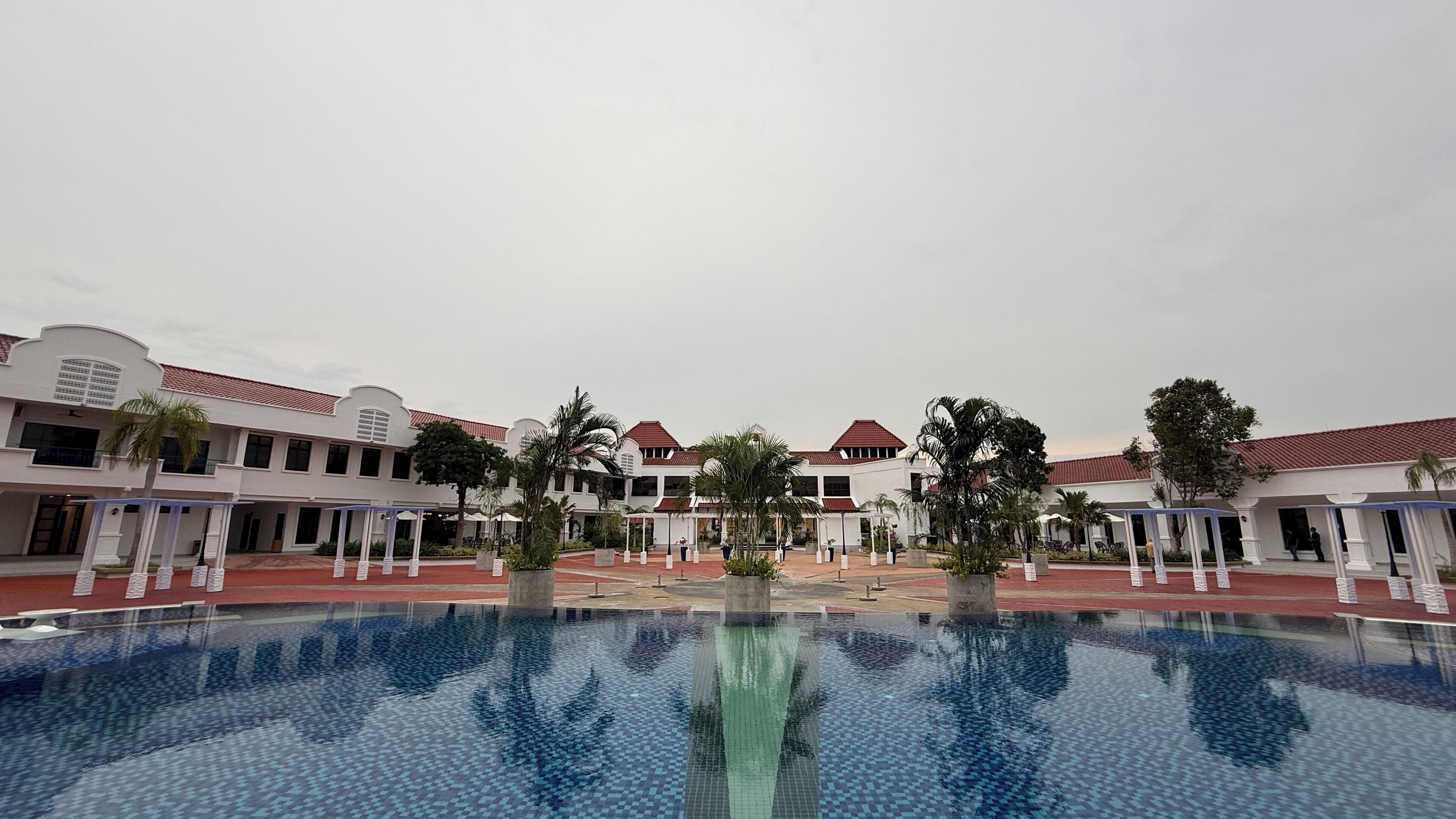 A hotel courtyard with a central pool, surrounded by white buildings with red roofs and palm trees.