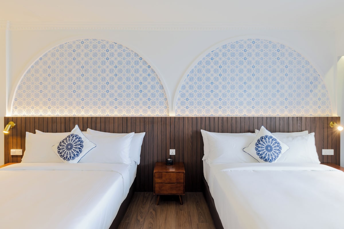 Une chambre d’hôtel avec deux lits, chacun recouvert d’une courtepointe blanche et agrémenté d’un coussin à motifs bleus. Une petite table de chevet en bois se trouve entre les deux lits, sur laquelle est posée une petite lampe.