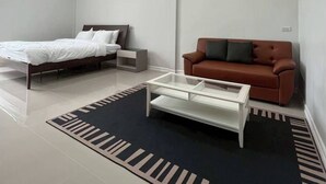 A modern bedroom with a bed, a brown sofa, a coffee table, and a patterned rug. - The Orange Grand Resort (Det Udom)