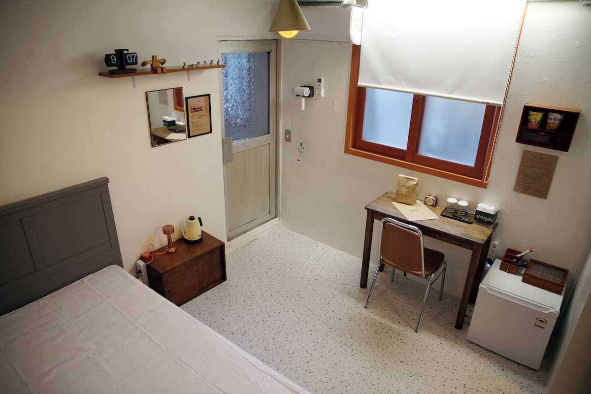 A small, well-lit room with a bed, a desk, a chair, and a small refrigerator.