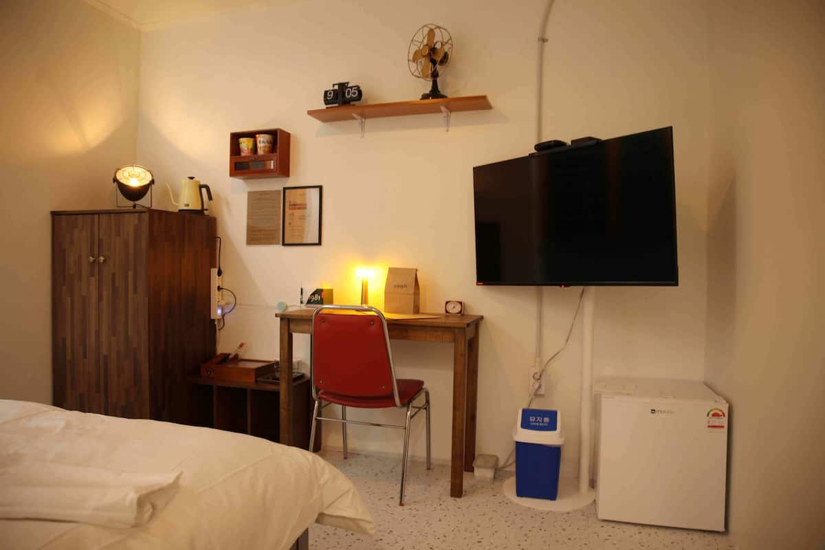 A bedroom with a bed, a desk with a chair, a television, a small refrigerator, and a wooden cabinet.