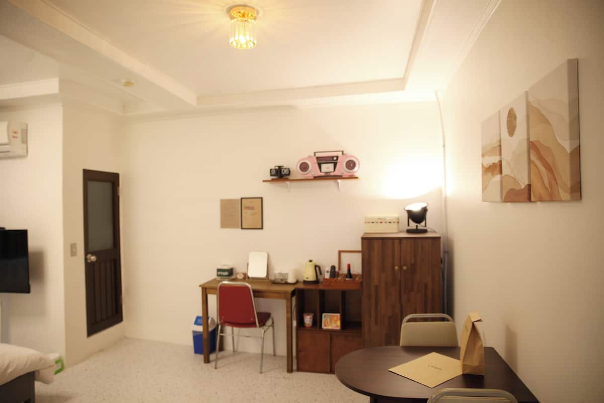 A small, well-lit room with a desk, chair, and a table. There is a radio on the shelf above the desk, a framed certificate on the wall, and a painting on the wall to the right.