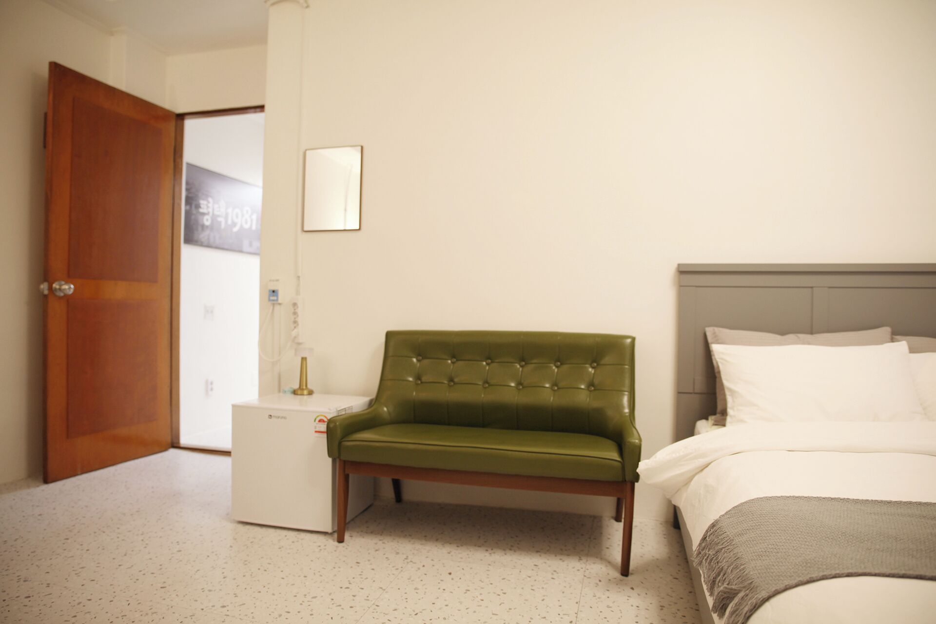 Una habitación de hotel con una butaca tapizada de verde, una cama con ropa blanca y una puerta de madera.