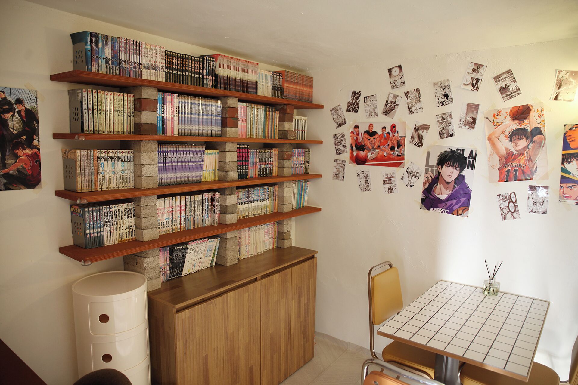 Una habitación con una estantería llena de libros, una mesita con mantel a cuadros y varios carteles en la pared.