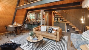 Un intérieur chaleureux et rustique, avec un plafond haut, des poutres en bois, un coin repas avec une table ronde, un canapé agrémenté de coussins à motifs et un escalier menant à un étage supérieur.
