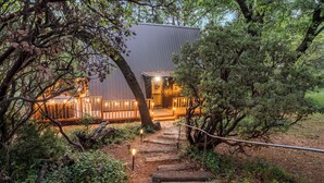 Un sentier en bois mène à un bâtiment à l’extérieur métallique, entouré d’arbres et illuminé par de petites lumières.