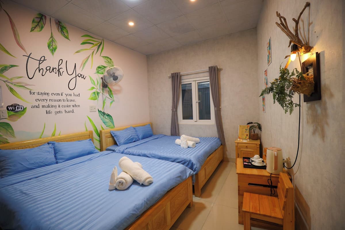 A hotel room with two beds, a small wooden table, a wall-mounted lamp, and a window with curtains.