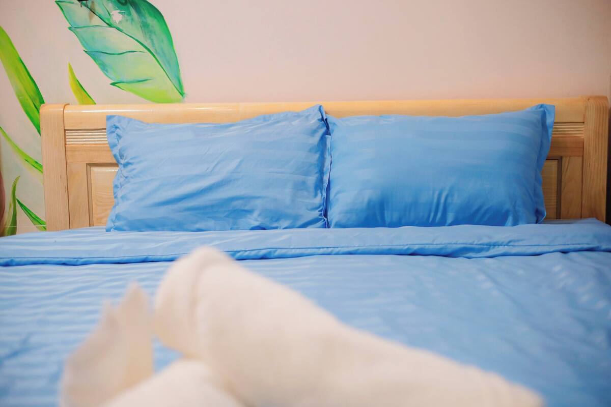 A bed with blue bedding and pillows, a wooden headboard, and a green plant to the left.