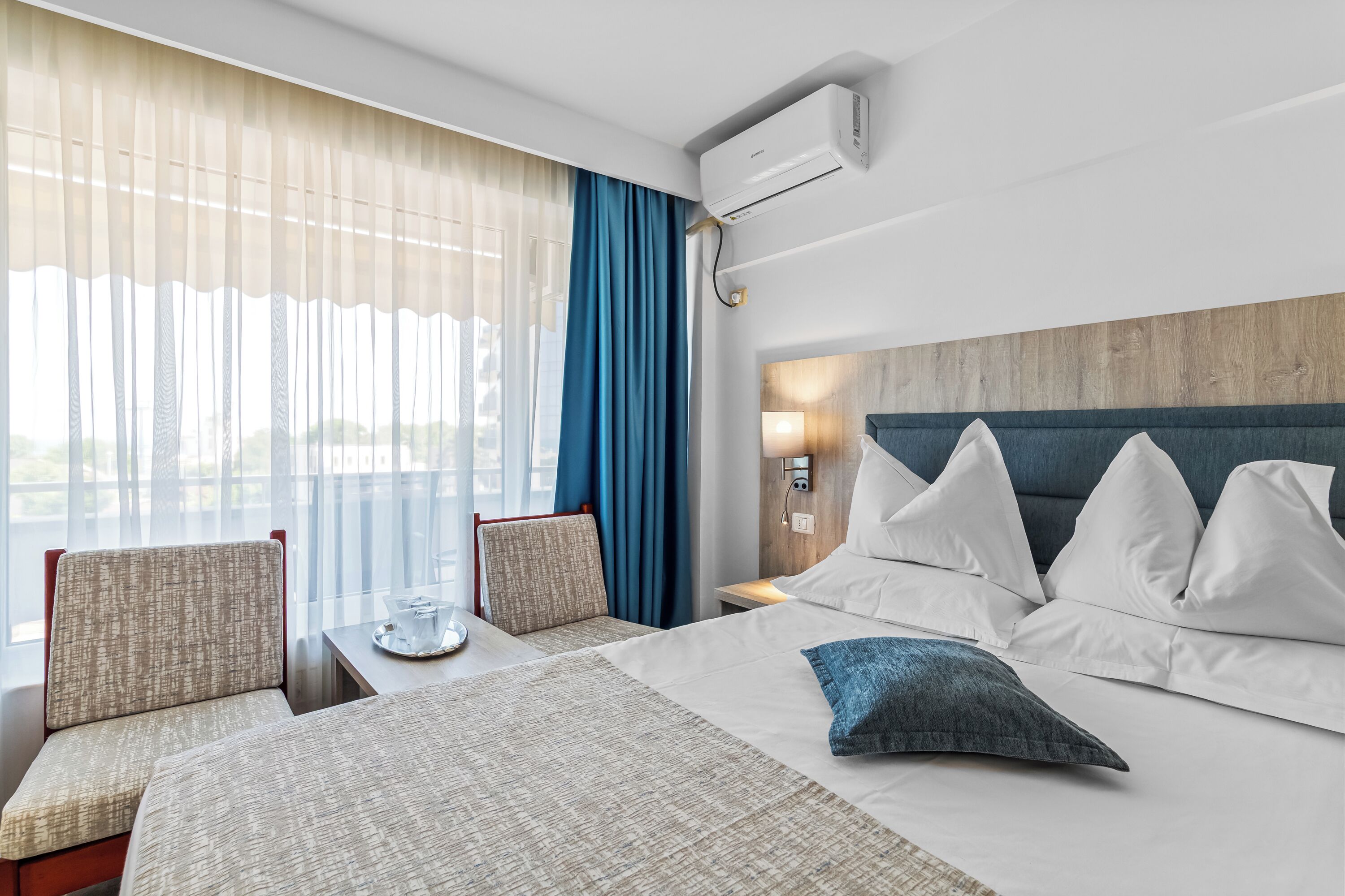 A hotel room with a bed, two chairs, a nightstand, and a window with curtains.