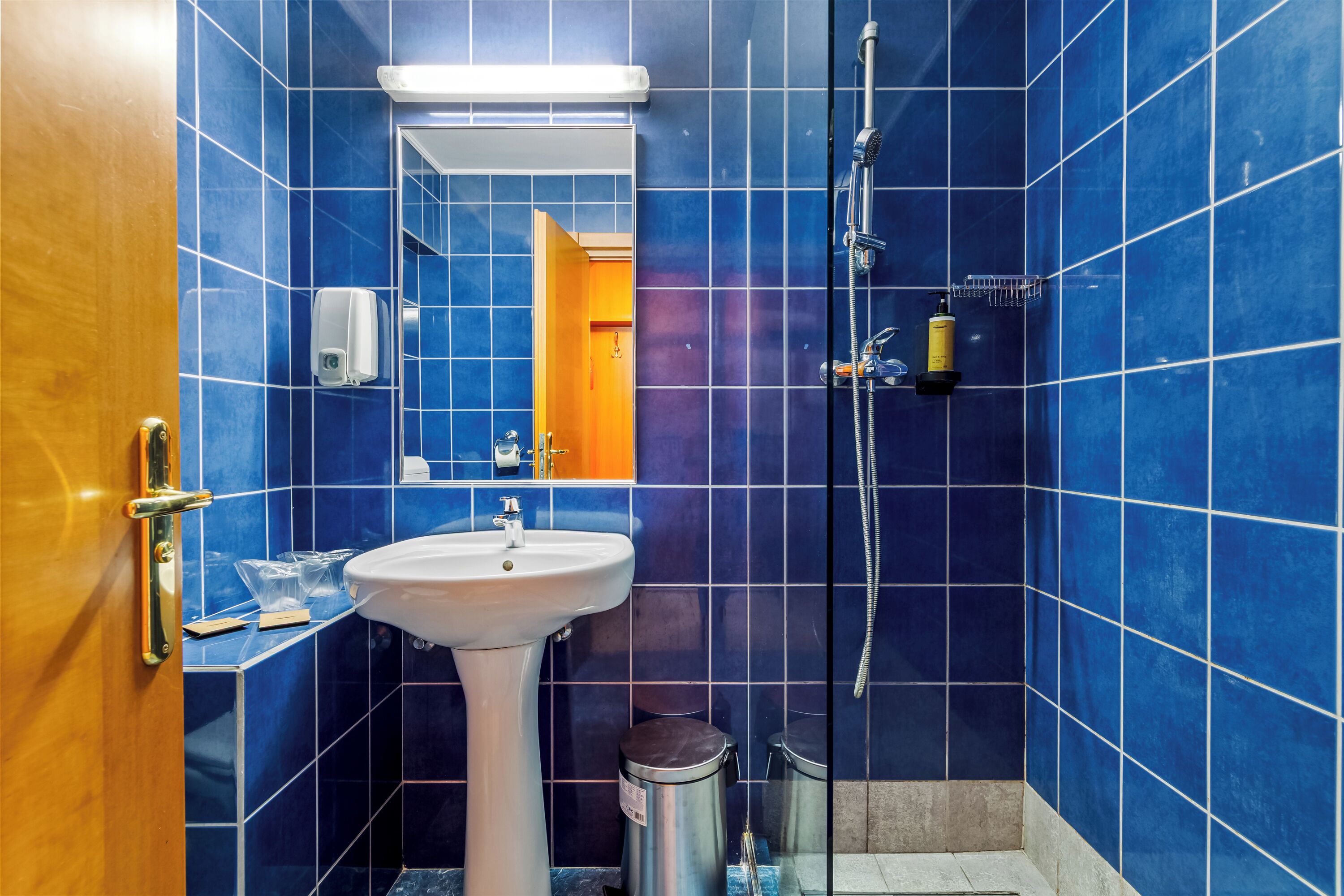 Een badkamer met blauwe tegels, een wastafel, een spiegel, een douche en een prullenbak.