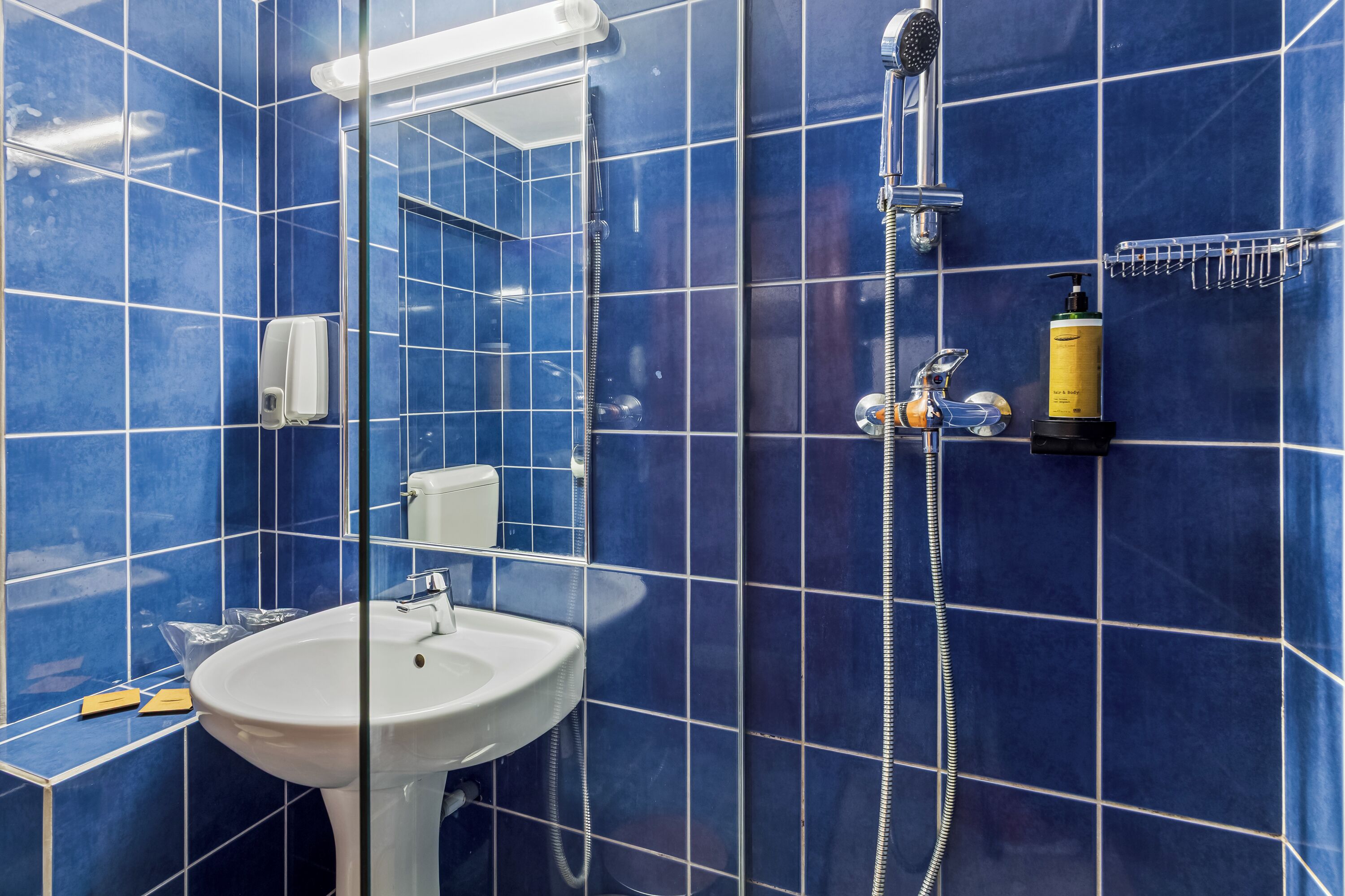 Un baño con azulejos azules, un lavamanos blanco, un cabezal de ducha de mano y un dispensador de jabón.