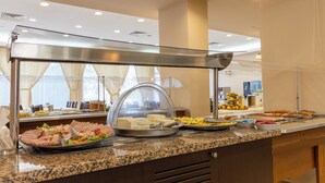 A buffet setup with various food items, including meats, fruits, and cheeses, in a well-lit room with a glass partition.