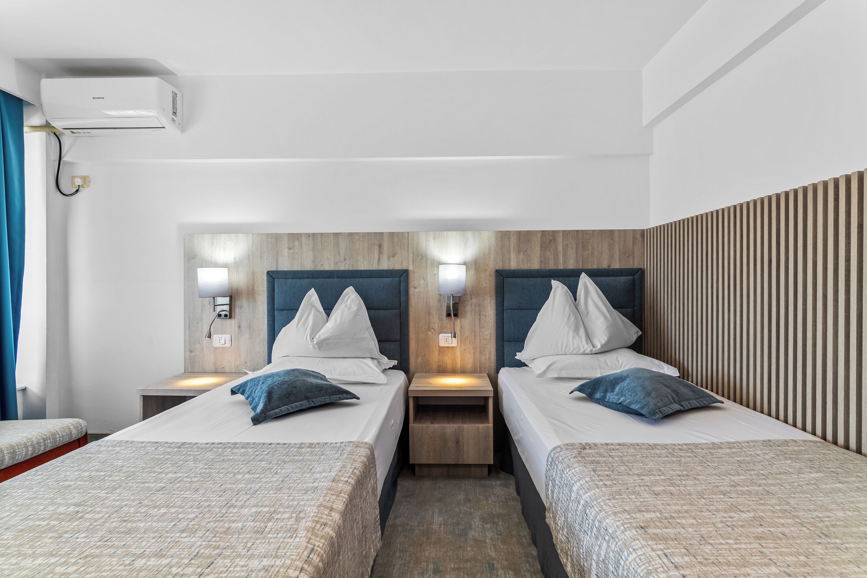 Habitación de hotel con dos camas, cabecero de madera, lámparas de noche y un equipo de aire acondicionado.